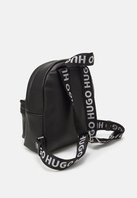 Mochila negra de cuero con forma redondeada, con una correa llamativa y de marca con el texto "HUGO" en blanco. Incluye detalle de bolsillo lateral.