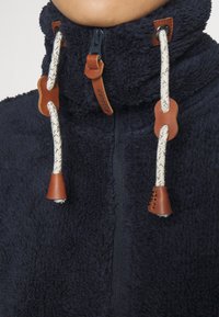 Giacca in pile navy con colletto alto, caratterizzata da dettagli in cuoio marrone e cordini con toggle. Tessuto morbido e particolari con chiusura a zip.