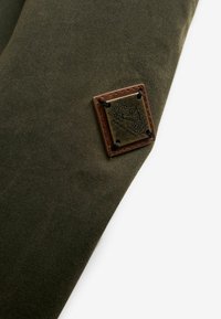 Olivově zelená látková nášivka s tlakovým vzorem a hnědými koženými akcenty; čtvercový tvar se zaoblenými rohy; upevněno černým šitím.