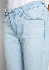 Jeans de ganga azul claro com um clássico design de cinco bolsos, fecho de botão metálico e um leve desbotado na textura do tecido.