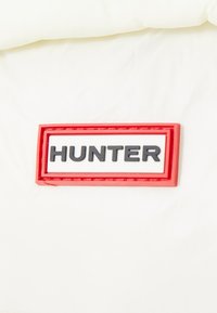 Etiqueta de tela blanca con texto en rojo y negro que dice "HUNTER". La etiqueta tiene forma rectangular y un borde texturizado.