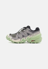Chaussure de trail de course grise avec une tige en mesh texturé, semelle en caoutchouc verte et des éléments de design distinctifs, y compris un système de laçage rapide et un logo.
