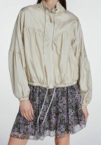 Giacca a vento beige chiaro con zip, colletto alto e vita regolabile con coulisse, con maniche arricciate, indossata sopra una gonna a stampa floreale.