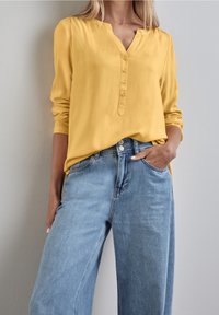 Gul långärmad blus med knäppning, kombinerad med ljusblå, vida denimjeans med framfickor och en något slitning.