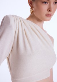 Haut asymétrique en tissu crème texturé avec un léger éclat, présentant des détails plissés sur la manche et un corsage ajusté.