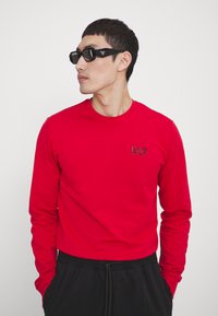 Maglietta rossa a maniche lunghe con scollatura a girocollo e logo EA7 sul petto, abbinata a occhiali da sole neri angolari e pantaloni neri.