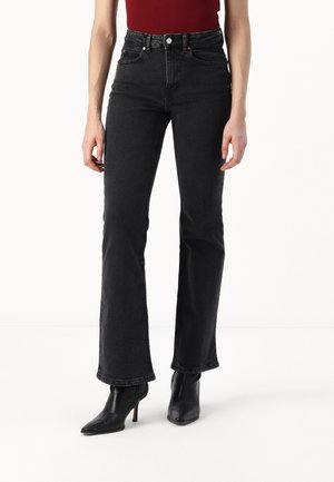 Kvinde iført sorte højtaljede, udvidede jeans, rød ærmeløs top og sorte ankelstøvler med spidse tæer og hæle, stående mod en hvid baggrund.