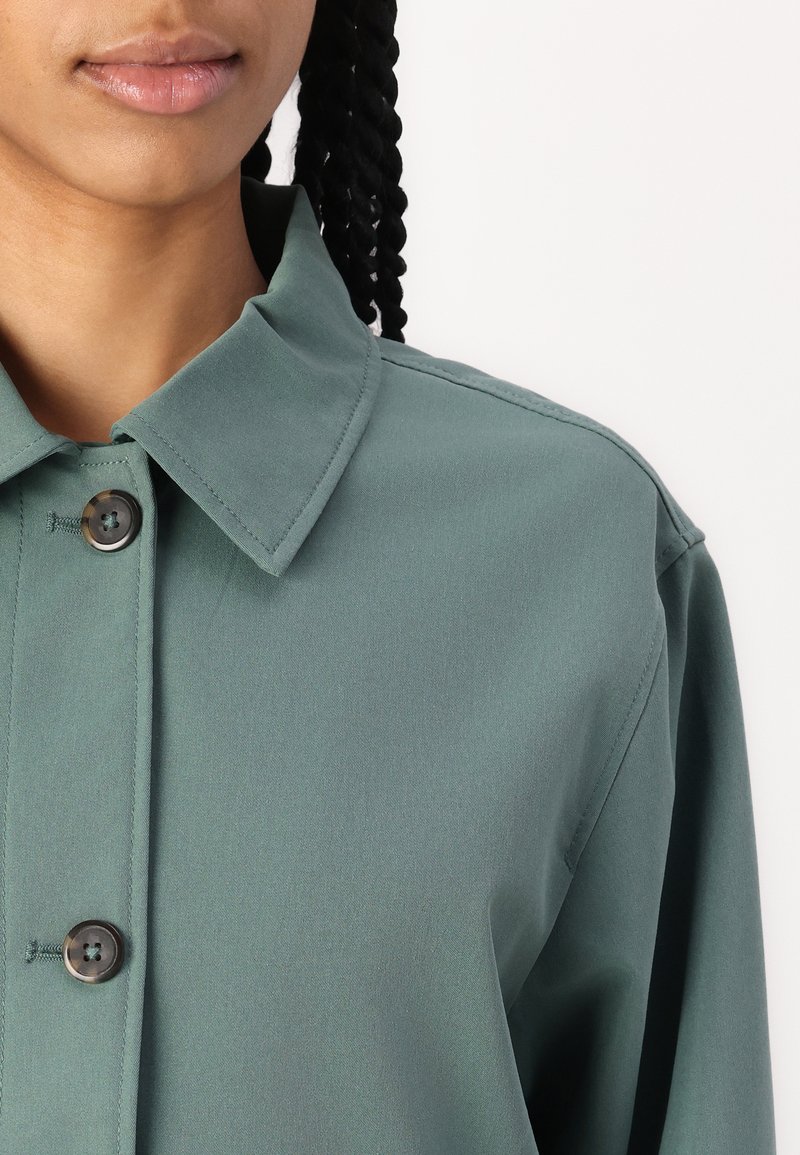 Groen overhemd met knopen en een op maat gemaakte kraag, met een subtiele glans. Bevat een donkere knop en een soepele stofstructuur. Geknipte weergave van het borstgebied.