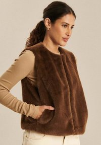 Kobieta z brązowymi włosami w kucyku, ubrana w bezrękawnik z brązowego sztucznego futra na beżowej koszulce z długim rękawem, z rękami w kieszeniach kamizelki.