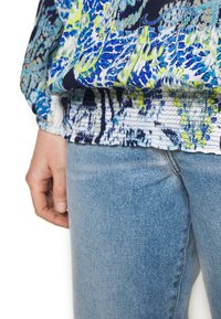 Maglietta lunga a maniche colorata e a fantasia con tonalità di blu, verde e bianco, caratterizzata da un orlo arricciato, abbinata a jeans azzurri chiari.