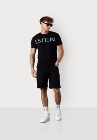 T-shirt noir avec un graphisme "ESTEEM" en multicolores, associé à un short noir et des baskets blanches. Modèle portant des lunettes de soleil. Tissu lisse.