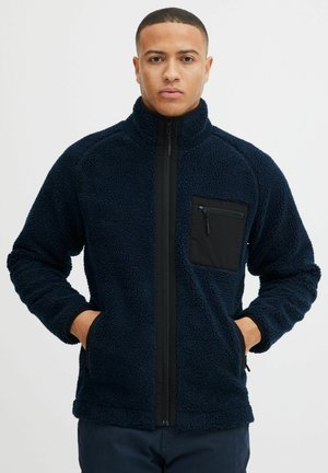 Veste en polaire marine avec une poche zippée noire, un col haut et un fermeture éclair à l'avant. Tissu texturé avec des poignets et un ourlet élastiques. Deux poches latérales.