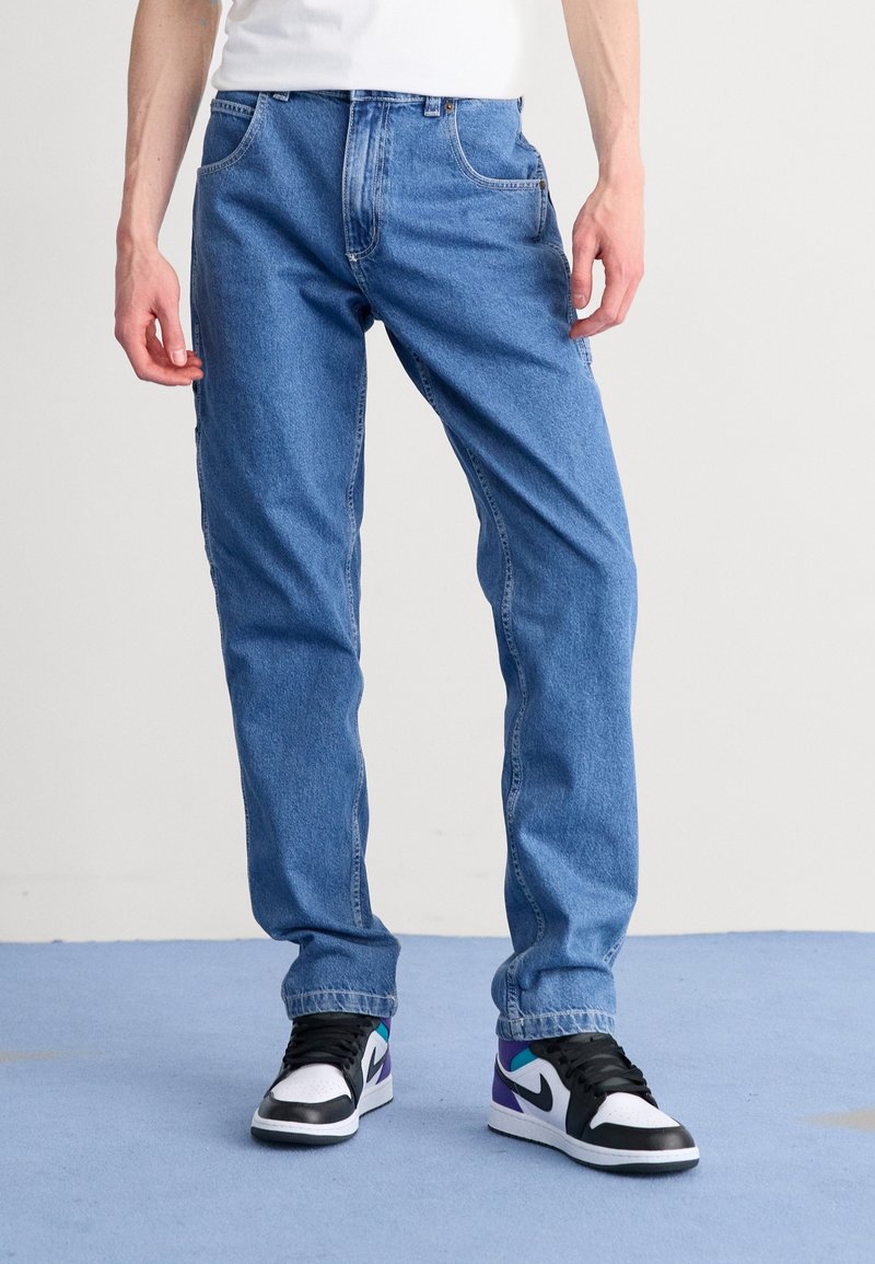 Personne portant un jean droit bleu et des baskets Nike noires, blanches, violettes et turquoise, debout sur un sol bleu.
