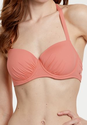 Kvinde iført en koralfarvet bikini med bøjle, rynket stof og halterneck-stropper, der viser overkroppen og hånden på hoften.