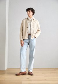 Beige Canvasjacke mit schwarzen Knöpfen, hellblaue Jeans und braune Lederschuhe. Das Outfit verfügt über einen Kragen und Taschen vorne.