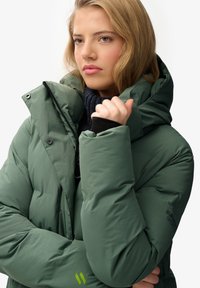 Grüne Pufferjacke mit hohem Kragen, gestepptem Muster und schwarzen Akzenten. Verfügt über ein Logo am Ärmel und eine Reißverschlusstasche.