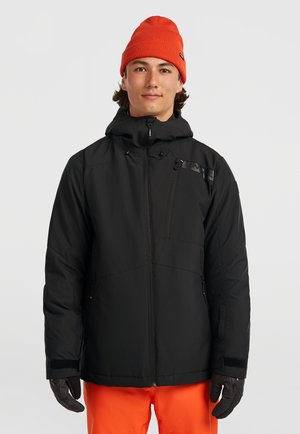 Zwarte pufferjack met een capuchon, voorzien van een ritssluiting, voorvak en contrasterende oranje beanie. Gekozen in combinatie met oranje broek en handschoenen.
