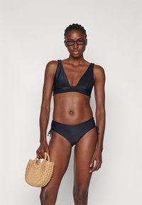 Set bikini nero a coste con top a profondo scollatura a V e slip con lacci laterali. La modella tiene una piccola borsa di paglia intrecciata. Texture liscia e aderente.