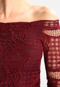 Top bez ramínek v burgundské barvě z krajky s květinovými a diamantovými vzory, odhalující holé rameno a poloprůhledné dlouhé rukávy.