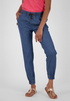 Personne portant un pantalon bleu avec poches élastiques et cordon de serrage, ainsi que des sandales en or rose, debout avec une main dans la poche.