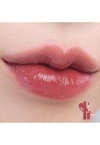 Glansig, dämpad rosa läppfärg på fylliga, släta läppar. I det nedersta hörnet syns en behållare för läppprodukt med en applikator.