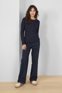 Námořnický modrý kardigan na knoflíky s kulatým výstřihem, spárovaný s odpovídajícími námořnickými kalhotami širokého střihu a béžovými teniskami s zelenými akcenty.