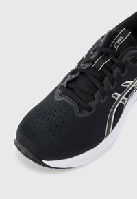 Chaussure de sport noire avec une tige en tricot et une semelle blanche. Les caractéristiques incluent un design à lacets, une surface texturée et un logo et des accents argentés contrastants.