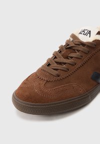 Sapatilha de camurça castanha com biqueira arredondada, superfície texturizada e logótipo preto contrastante. Apresenta atacadores planos e sola de borracha.