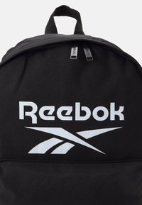 Črn nahrbtnik iz tkanine z belim logotipom in grafiko "Reebok". Okrogla zgornja stran, dvojno zadrgo in zgornji ročaj za prenašanje.