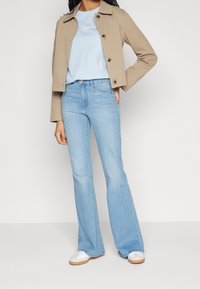 Beige lyhyt takki napilla, vaaleansininen t-paita ja väljät vaaleansiniset farkut. Valkoiset lenkkarit vaaleanruskeilla yksityiskohdilla. Yksinkertainen muotoilu.