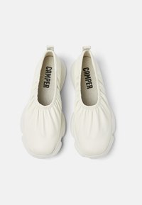 Scarpe bianche senza lacci con bordi arricciati, in morbido materiale di pelle e suola imbottita. Presentano il nome del marchio "CAMPER" ben visibile all'interno.