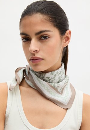 Junge Frau mit zurückgebundenen dunklen Haaren, trägt ein weißes Tanktop und einen gemusterten beigen und grünen Schal, der um ihren Hals gebunden ist.