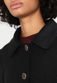 Cappotto nero con tessuto strutturato, caratterizzato da un colletto arrotondato e bottoni neri con dettagli dorati sulla parte anteriore.