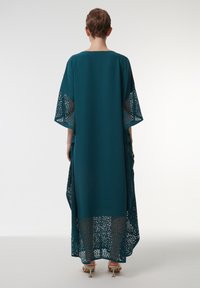 Vestido largo en tono verde azulado con un corte holgado, que presenta intrincados diseños cortados a láser en las mangas y el dobladillo. Textura de tejido suave y mangas amplias.