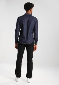 Tmavě modrá džínová košile s dlouhými rukávy, špičatým límečkem a detailem zadního dílu, doplněná o černé kalhoty a bílé tenisky.