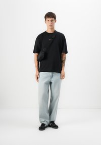 Young man wearing black oversized t-shirt, light blue wide-leg jeans, black sneakers, and black crossbody bag standing against white background.