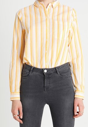 Femme portant une chemise à rayures verticales jaunes et blanches, boutonnée, rentrée dans un jean taille haute gris foncé.