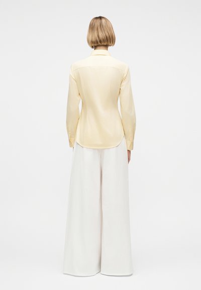 Άτομο με κοντά ξανθά μαλλιά φοράει ανοιχτό κίτρινο πουκάμισο με μακριά μανίκια και λευκό παντελόνι με φαρδιά μπατζάκια, στέκεται γυρισμένο με την πλάτη προς το απλό φόντο.