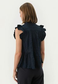 Dunkelblausa Bluse aus strukturiertem Stoff, mit rüschierten Schultern und einem gestuften Saum, hohem Halsausschnitt und lockerer Passform.