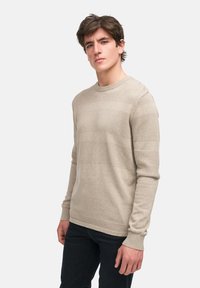 Beige Strickpullover mit strukturiertem Muster und gerippten Manschetten. Rundhalsausschnitt und lässiger Schnitt. Kombiniert mit dunklen Hosen.