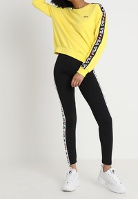 Sudadera amarilla recortada con logo en el lado izquierdo del pecho, combinada con leggings negros con rayas laterales y zapatillas blancas.