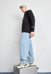 Felpa nera con grafica frontale, abbinata a jeans baggy blu chiaro e sneaker beige con strisce nere e bianche, in piedi su una scatola metallica.