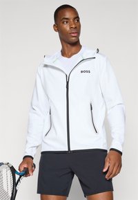 Weiße Zip-Jacke mit Kapuze, ausgestattet mit schwarzen Akzenten, zwei Seitentaschen und einem Logo auf der Brust, getragen über einem weißen T-Shirt und schwarzen Shorts.