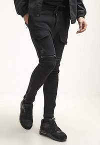 Svarta joggerbyxor med slim fit, med cargofickor, dragkedjedetaljer, ribbad textur och en midja med snörning. Ett par svarta sneakers syns.