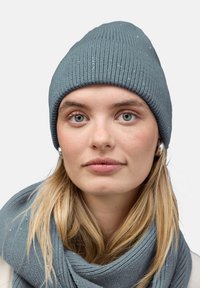 Gorro de punto en color teal con textura acanalada y lentejuelas dispersas; combinado con una bufanda a juego en un estilo similar, colgada alrededor del cuello.