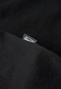 Tessuto di cotone nero per t-shirt con un'etichetta cucita visibile che recita "Superdry". Texture liscia con un design semplice e senza motivi.