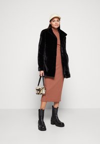 Manteau en fausse fourrure noire, robe moulante marron, bottines noires à semelles épaisses et sac à main à motifs. Casquette de couleur claire sur la tête.