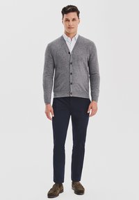 Mężczyzna stoi, ubrany w szary kardigan zapinany na guziki, biały koszulę w wzory, granatowe spodnie i brązowe mokasyny, na tle gładkiego białego tła.