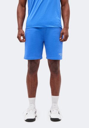 Mand iført blå sportsshorts og matchende trøje, hvide crew-sokker og hvide Adidas-sneakers, stående mod en ensfarvet lys baggrund.