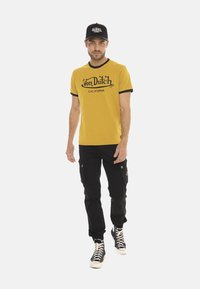 T-shirt jaune avec des détails en noir à l'encolure et imprimé "Von Dutch CALIFORNIA". Associée à un pantalon cargo noir et une casquette noire, en position de marche, baskets.